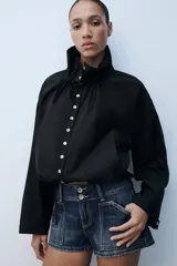 Cazadora de algodón negra con cuello subido ajustable con lazadas, manga larga, bolsillos de vivo en los laterales y bajo ajustable con cordones. Cierre frontal con botones.