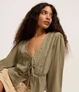 Blusa tipo bata de algodón color verde oliva, con escote en V, cierre frontal mediante lazos, mangas largas y detalles de bordados florales y encaje en el frente.