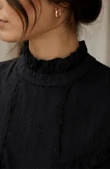 Blusa negra de tela con textura calada, cuello alto con volado, y detalles de volados verticales en el frente.