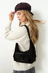 Cartera de hombro de símil gamuza color negro, con diseño de media luna, bolsillo frontal con cierre y bolsillos laterales. Cuenta con correa ajustable con ojales metálicos.