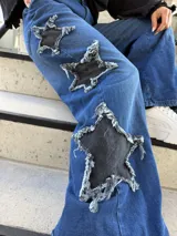 Pantalón jeans azul de corte wide leg, tiro medio y diseño holgado, con grandes aplicaciones de estrellas negras desgastadas en los laterales de las piernas.
