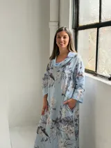 Vestido largo celeste con estampado floral en tonos blancos y grises, de corte holgado, mangas largas y cuello drapeado.