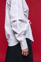 Camisa blanca de manga larga con volumen y estructura, cierre frontal con botones y lazo ajustable en la cintura que realza la silueta.