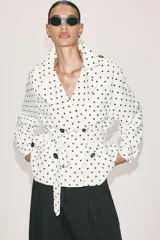 Cazadora estilo trench corta confeccionada en lino, con diseño de lunares negros sobre fondo blanco. Cuenta con cuello solapa, manga larga con trabillas en los puños, cierre frontal cruzado con botones negros y cinturón ajustable del mismo tejido.