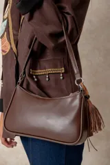 Cartera tipo baguette de diseño minimalista, confeccionada en una combinación de gamuza color beige en el panel frontal y cuero color chocolate en los bordes y la correa. Incluye un detalle colgante de cuentas de madera y cierre superior.