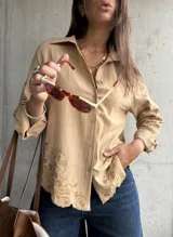 Camisa de lino color camel con cuello clásico, cierre frontal de botones y mangas largas. Presenta un delicado bordado floral tono sobre tono en las mangas y en el ruedo, el cual tiene un acabado festoneado.