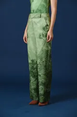 Pantalón de corte recto y tiro alto, de color beige con un estampado paisajístico de árboles y naturaleza en tonos marrones y grises. Cuenta con trabillas en la cintura y cierre frontal oculto.