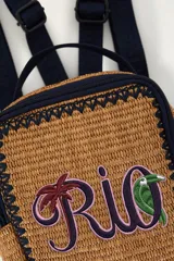 Bolso tote de mano color tostado, tejido con textura tipo mimbre o rafia, con bordes y detalles en azul marino. Cuenta con un asa superior corta, cierre con cremallera y un bordado frontal con la palabra "Rio" que incluye motivos tropicales de una palmera y un tucán.