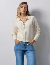 Cardigan de tejido punto color crema, con cuello tipo solapa, cierre frontal completo con tirador circular metálico y dos bolsillos de parche simulados en el frente. Presenta detalles de canalé en los hombros.