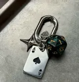 Llavero metálico plateado con mosquetón, que incluye varios dijes: una placa rectangular con el As de Picas, un dado poliédrico verde oscuro y dorado, una estrella de cuatro puntas, y un dije pequeño con forma de corazón que dice "made with love".