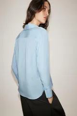 Camisa de cuello solapa y escote en pico, con manga larga y cierre frontal mediante botones. Presenta un diseño fluido con bajo asimétrico y aberturas laterales.