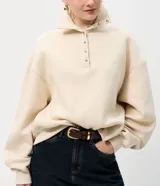 Buzo de algodón color beige, con cuello polo y cierre frontal parcial con botones dorados. Presenta un diseño de corte holgado con mangas largas y puños acanalados.