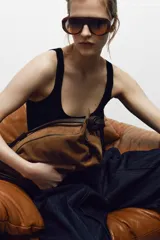Bolso de mano formato alargado confeccionado en piel, con doble asa, una de mano y otra de hombro, con detalle de nudos y ojales metálicos. Cuenta con cierre mediante solapa y cremallera, y bolsillo interior con cremallera.
