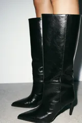 Bota de caña alta y ancha confeccionada en piel, con diseño de punta fina y tacón alto tipo aguja. Presenta costuras decorativas a lo largo del cuerpo y un diseño de calce fácil.