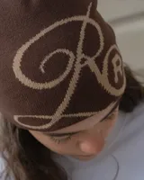Gorro tipo beanie tejido en color marrón oscuro con diseño gráfico de letras entrelazadas en color beige.