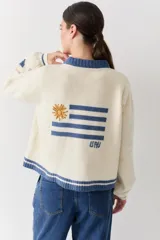 Cardigan de lana color crema con cuello redondo, cierre frontal de botones y mangas largas con puños ajustados. Presenta bordados decorativos en el frente que incluyen un sol, olas y estrellas, con detalles en azul en el cuello y el ruedo.
