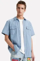 Camisa de manga corta estilo western, confeccionada en tejido chambray de algodón color celeste. Presenta cuello clásico, cierre frontal con botones a presión, dos bolsillos frontales con solapa y etiqueta roja distintiva de la marca.
