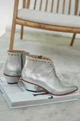 Botín plateado de cuero con taco bajo y punta fina. Presenta tachas alrededor del tobillo y cierre lateral.