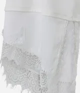 Remera de algodón corte oversized, color blanco, con cuello redondo, mangas cortas amplias y ruedo asimétrico con detalle de encaje aplicado.