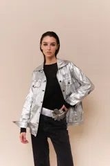 Chaqueta de cuerina metalizada color plateado, con corte oversized, cuello camisero y cierre frontal con botones. Presenta cuatro bolsillos frontales con solapa y botón, y logo bordado en las mangas.