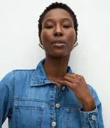 Vestido midi de denim con cuello camisero, mangas largas con puños abotonados y cierre frontal completo con botones metálicos. Presenta bolsillos aplicados en el pecho y un corte recto estructurado.