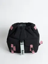 Mochila acolchada de nylon color negro con correas y detalles en rosa pálido. Presenta un cierre principal tipo solapa con hebilla de plástico negra y una cinta colgante con el texto "rusty" en color verde menta.