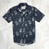 Camisa de hombre de manga corta con estampado floral en tonos grises sobre fondo azul marino. Tiene cuello clásico, cierre frontal con botones blancos y corte recto.