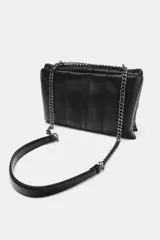 Bolso bandolera de diseño acolchado en color negro, con solapa frontal decorada con una cadena metálica y charms colgantes. Cuenta con cierre de botón imantado, triple compartimento interior y correa ajustable que permite llevarlo al hombro o cruzado.
