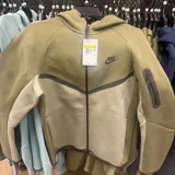 Campera deportiva Nike Tech Fleece con cierre frontal, capucha y bolsillo con cierre en la manga. Presenta un diseño de paneles en tonos verde oliva y beige, con el logo de la marca estampado en el pecho.