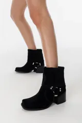 Bota de caña media confeccionada en cuero gamuzado color chocolate. Presenta un diseño de estilo western con taco cuadrado de 7 cm y una correa decorativa en el tobillo con herrajes metálicos plateados.