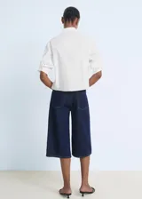 Camisa de corte recto y diseño crop, confeccionada en popelín de algodón. Presenta cuello camisero, cierre frontal con botones, mangas tres cuartos y un bolsillo en el pecho con detalle de strass.