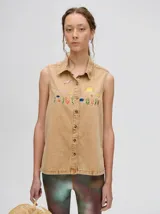 Camisa musculosa de denim liviano en color tostado, con cuello camisero y cierre frontal de botones. Presenta bordados coloridos en el pecho con motivos veraniegos como el sol, olas y peces, además de un detalle bordado en el cuello.