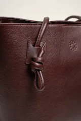 Cartera tote de cuero suave color marrón, con diseño amplio y líneas simples, con tiras de hombro decoradas con nudos artesanales y pequeño logo grabado en el centro.