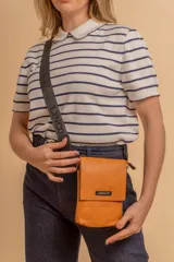 Phonebag pequeña color mostaza, confeccionada en cuero sintético, con solapa frontal y correa ajustable con logo de la marca.