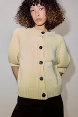 Chaqueta de punto color amarillo claro, con cuello redondo, mangas abullonadas por debajo del codo y cierre frontal con botones oscuros.