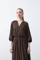 Vestido largo color marrón, de silueta fluida, con escote en V y botones frontales. Presenta cintura elastizada y mangas largas con elástico en los puños.