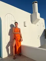 Vestido largo color naranja, con escote halter cruzado y falda tipo pareo con tajo lateral. Confeccionado en tela crepé con caída fluida.