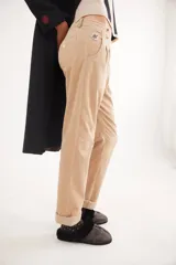 Pantalón recto de tiro medio confeccionado en pana con lycra. Presenta pinzas frontales, bolsillos laterales y traseros, y cierre con botón dorado. El diseño destaca por el detalle de paño espigado en el interior del ruedo, visible al doblarlo, y un pin bordado decorativo en el bolsillo delantero.