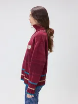 Pullover de textura tipo terciopelo con cuello alto y cierre frontal parcial. Presenta un diseño a rayas horizontales en tonos grises y negros, con detalles en rojo y blanco en el cuello, puños y ruedo. Incluye un aplique bordado con la letra M en el pecho y puños acanalados.