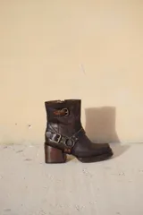 Bota corta de cuero en tono marrón oscuro con efecto desgastado, taco cuadrado de madera y punta cuadrada. Presenta correas decorativas con hebillas metálicas y herrajes en los laterales.