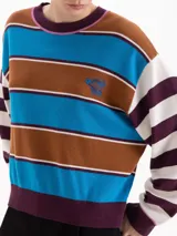 Sweater de punto con diseño de rayas horizontales en tonos marrón, azul, blanco y bordó. Presenta cuello redondo, mangas largas y un pequeño bordado en el pecho. Su corte es holgado y de largo a la segunda cadera.