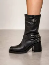 Bota biker de cuero negro con caña media, punta cuadrada y taco cuadrado. Presenta detalles de correas con hebillas metálicas en el tobillo y en la parte superior de la caña.