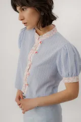 Blusa celeste a rayas blancas, de manga corta abullonada. Cuenta con cuello mao y tapeta de botones con detalles de estampado floreado en tono rosado, presentes también en el cuello y los puños.