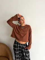 Remera de manga larga color marrón, con cuello redondo y detalle de parche cuadrado con logo bordado en el centro del pecho. Presenta un corte holgado y diseño minimalista.