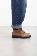 Borcego de cuero vacuno color marrón caramelo con acabado mate, estilo combate y caña baja. Cuenta con cordones marrones, panel de gamuza oscura en el cuello y suela de caucho negra tipo tractor con costura blanca visible.
