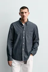 Camisa de corte relajado confeccionada en tejido de algodón denim, con cuello solapa, manga larga con puños abotonados y bolsillo plastrón en el pecho. Presenta cierre frontal de botones y bajo con aberturas laterales.