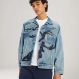 Chaqueta de denim de corte holgado, lavado medio, con bolsillos frontales con solapa, cierre de botones metálicos y detalles de parches e impresiones en tonos más oscuros.