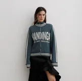 Campera tejida de algodón en color rojo, con cierre frontal completo, cuello alto y puños acanalados con detalles de rayas blancas. Presenta un diseño gráfico con texto blanco en el frente.