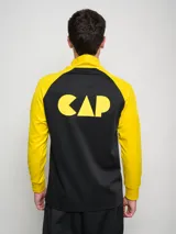 Buzo deportivo de entrenamiento, color negro en el torso y amarillo en las mangas y cuello. Es de manga larga, con cuello alto y cierre parcial frontal. Presenta un escudo con estrellas en el pecho.