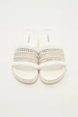Sandalias de base plana color marfil con tira ancha decorada con bandas de cordón trenzado, detalles plateados con tachas y flecos en los bordes.